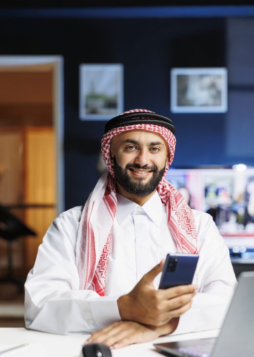 Male Arab person in profile, grinning at camera with his personal computer close by. Using contemporary technology, smiling Muslim guy with a cell phone, portrayed in portrait with laptop on table.